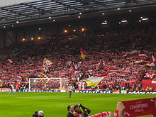 A stand which is full of people standing to support their team. There are a number of flags, scarfs, and banners in the crowd.