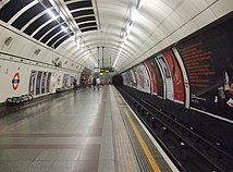 Angel tube station platforms. There roof is shaped in an arc, and London Underground roundels - red circles with dark blue lines through them - are on the walls.