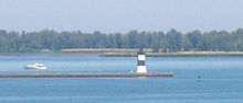 A pier with a square, black and white-striped lighthouse at the end. A small motorboat is paralleling the pier, heading away from the lighthouse.