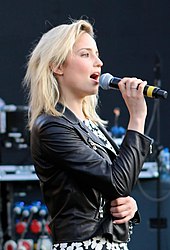 A blonde woman in a leather jacket and floral dress holding a microphone.