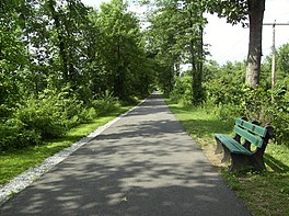 A photograph of the rail trail, with grass on either side. A green park bench is to the right of the trail and various trees are in the distance.