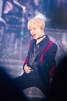 Jimin stands on a stage holding his belt while dancing.