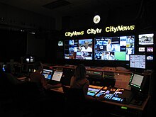 A darkened television control room. Above a bank of monitor displays are a backlit clock and lit signs for CityNews and Citytv. Several people are seated at switchers and computers, controlling the broadcast.