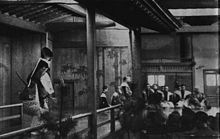 Photo noir et blanc de 1943. Au premier plan à gauche, l'acteur s'avance sur le pont ; à droite, les spectateurs vus de dos. Au fond à gauche, la paroi est ornée de bambous stylisés, à droite, musiciens accroupis sur la scène.