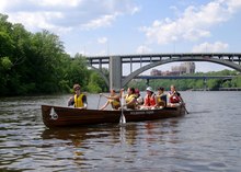 Seven young people in canoe, shoreline is green, women paddling, all wearing life vests, bridge span and university visible behind them