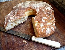 La photographie représente une couronne de pain. Le quart enlevé montre une mie aux bulles irrégulières sous une croûte dorée et farinée. Le pain repose sur une planche à pain en bois avec un couteau à dents à manche de plastique blanc.