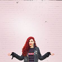 A young woman with curly red hair wearing a jacket stands in front of a pink-painted brick wall with her arms partially outstretched and her palms facing upward.