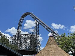Appearing from the central plaza, Iron Gwazi's lift hill is prominently shown in the skyline with the barrel roll-down drop under the lift structure. Parts of the building that houses the hybrid roller coaster are imaged at the bottom, with a thatched roof in the center-right. Trees are also visible on either side of the image.