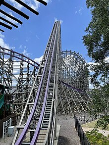 The Iron Gwazi lift hill is depicted from the queue line bridge that sits above where the train initially descends into the lift. The purple track significantly appears into the skyline. Parts of the barrel roll downdrop under the lift hill and the exit from the wave turn can be observed.