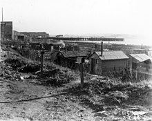 Row of shanties in a Hooverville with smoke coming out of several chimneys. The Alhambra Stucco Company is on the left side of image.