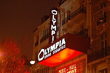 Photographie en couleurs représentant la façade d'une salle de concerts, de nuit, dans une lumière de néons orangés.