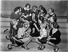 Photographie en noir et blanc représentant, sur des marches, Jacques Brel assis au centre, entouré d'une dizaine de jeunes femmes toutes tournées vers lui.