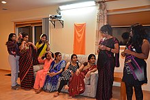 A group of young women sit and stand around a sofa as they finish preparing their hair and outfits.