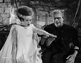 The Bride of Frankenstein has black hair with a white streak running through it, is dressed in a white gown, and has a blank expression. She is standing on the left with her left hand elevated. On the right is Frankenstein's monster, standing on the right and smiling. His right hand is below hers. The background includes walls made of stone.