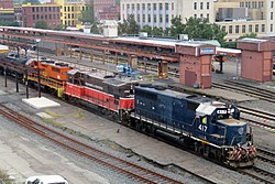 A freight train passing through a train station, lead by three diesel locomotives in various paint schemes.