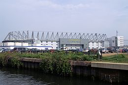 The exterior of Carrow Road, an association football stadium. A river is in the foreground.