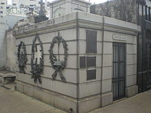 Photograph showing a mausoleum building of gray ashlar construction with metal wreaths and plaques attached to the outside walls, a metal barred gate at the entrance and surmounted by a square cupola