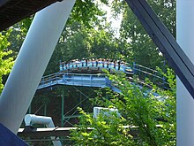 A SooperDooperLooper train is depicted as cresting one of its various hills with passengers aboard. Trees and foliage cover the background and foreground of the roller coaster.