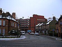 A tall red brick building towers over streets of two-storey houses. The roofs of the houses and the surrounding pavements are covered in snow.