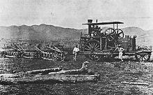 Photographie d'un imposant tracteur à vapeur dans un champ avec des collines à l'arrière-plan. Trois hommes se tiennent autour du véhicule.