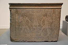 Large cubic block of granite decorated with figures, one of which has its arms raised up towards the sky.