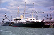 Her Majesty's Yacht Britannia is docked in Portsmouth Harbour for the 50th anniversary of the D-Day Landings in 1994. More modern Royal Navy ships are docked in behind her, and the masts of the HMS Victory can be seen in the far background.