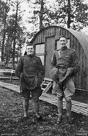 Two men in military uniform standing side-by-side. The man on the left is short and stocky, and the man on the right is tall and thin. In the background is a cabin.