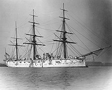 Starboard broadside view of a metal sailing ship in port at anchor. The three masts are rigged with yards and furled sails. A smokestack is amidships and ventilators rise above the top deck. Guns are visible protruding through the sides of the ship.