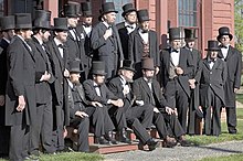 Seventeen men dressed as Abraham Lincoln, in black, with beards, and stovepipe hats.