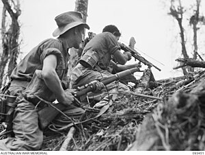 Two soldiers crouching on an incline in jungle terrain. The man on the left is holding a rifle and the man on the right is firing a light machine gun