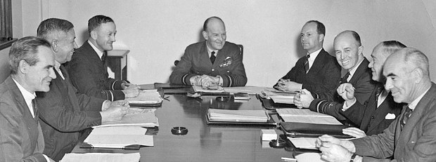Eight men seated at a table, three in military uniforms and the others in business suits