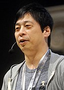 A black-haired Japanese man in his fifties at a convention, wearing a light grey t-shirt and paler grey hoodie.