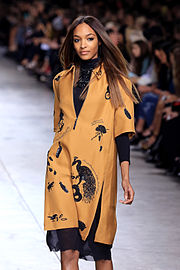 A brunette woman walks down a fashion runway in a tan-colored zipper garment over a black turtleneck dress