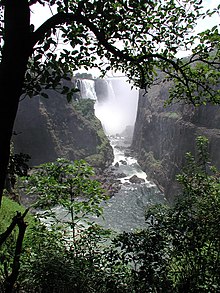 A majestic African scene. The viewer stands beneath a tree, overlooking the a large river from a high vantage point on a pleasant, sunny day. On each side of the river a great cliff rises above the viewer. In the distance an enormous waterfall can be seen, the spray from the water obscuring much of the view. The land is green and lush, and the river shimmers in the sun.