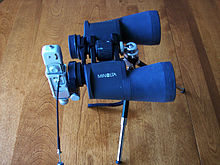 Small, spy sized camera attached to one eyepiece of a set of prismatic binoculars mounted to a tripod on a table top
