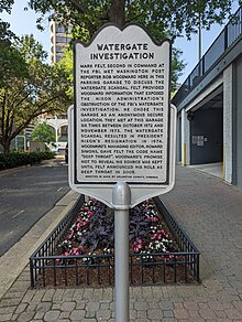 Photo of a grey historical marker titled "Watergate Investigation" on the sidewalk beside a small urban street with a parking garage door visible on the right behind the sign. The marker reads: "Mark Felt, second in command at the FBI, met Washington Post reporter Bob Woodward here in this parking garage to discuss the Watergate scandal. Felt provided Woodward information that expose the Nixon Administration's obstruction of the FBI's Watergate investigation. He chose this garage as an anonymous secure location. They met at this garage six times between October 1972 and November 1973. The Watergate scandal resulted in President Nixon's resignation in 1974. Woodward's managing editor, Howard Simons, gave Felt the code name 'Deep Throat'. Woodward's promise not to reveal his source was kept until Felt announced his role as Deep Throat in 2005. Erected in 2008 by Arlington County, Virginia."