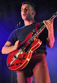 A man wearing skinny jeans and a dark t-shirt performs music with an electric guitar.