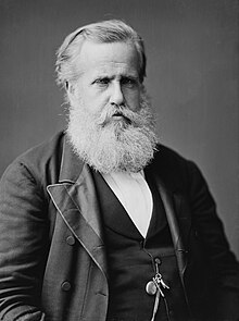 Hauf-length photographic portrait o an aulder man wi white hair an beard dressed in a dark jacket an necktie