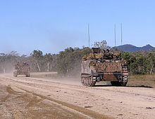 Colour photo of two tracked military vehicles driving on a dirt road