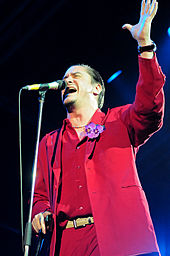 A man in a red suit standing in front of a microphone stand, with one arm raised