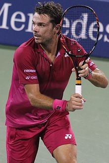 A man wearing a red shirt and red shorts grips his tennis racket in front of him as he stares to his right