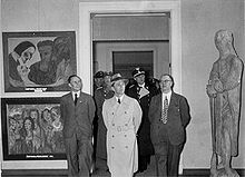 Frontale Schwarzweißfotografie von einer Gruppe, die durch eine Tür in die Ausstellung geht. Vorne begleiten zwei Männer mit Anzügen Joseph Goebbels, denen Uniformierte folgen. Links sind zwei Gemälden und rechts eine Steinfigur zu sehen.