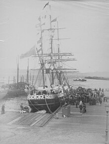 A crowd of people stand on a dock in front of a three-masted tall ship. The sails are down but there are many flags flying.