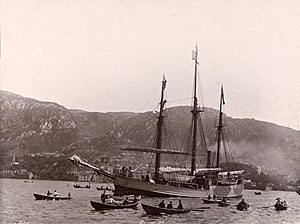 A three-masted ship, under steam power, moves across a stretch of water attended by several rowing boats. In the background is a line of hills, with buildings faintly visible at the water's edge.