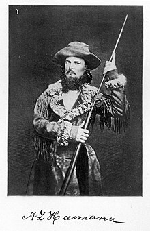 Three quarter length photo of Heermann holding a rifle and wearing a buckskin hat and a fur-lined coat. Heerman has a long, curly, black beard and long mustache.