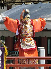 Danseur revêtu d'un demi-masque de chat, costume orange, pantalon et tunique à motifs floraux vifs.