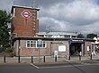 A red-bricked building with a rectangular, dark blue sign reading "REDBRIDGE STATION" in white letters all under a dark blue sky with white clouds