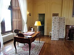 Photographie en couleurs d'une meuble en bois décoré sur lequel sont posés, des accessoires : lampe de bureau, sous-main, registres.