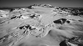 A snow-covered plateau with four small cone-shaped mounds in the foreground and a much larger flat-topped mountain in the background