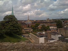 A scene of rooftops; on the left, a grassed mound with a tree; a rough stone wall in the foreground